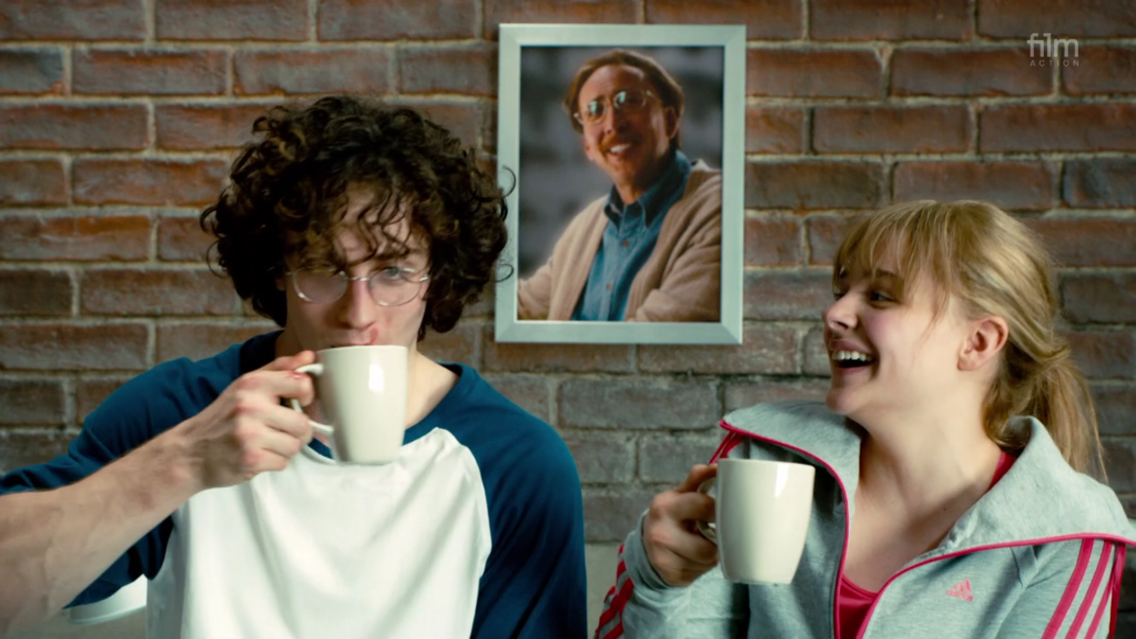 Chloë Grace Moretz as Hit Girl in Kick Ass 2, having a drink and laughing with the character Dave / Kick Ass, in front of a photo of Hit Girl's father Big Daddy.