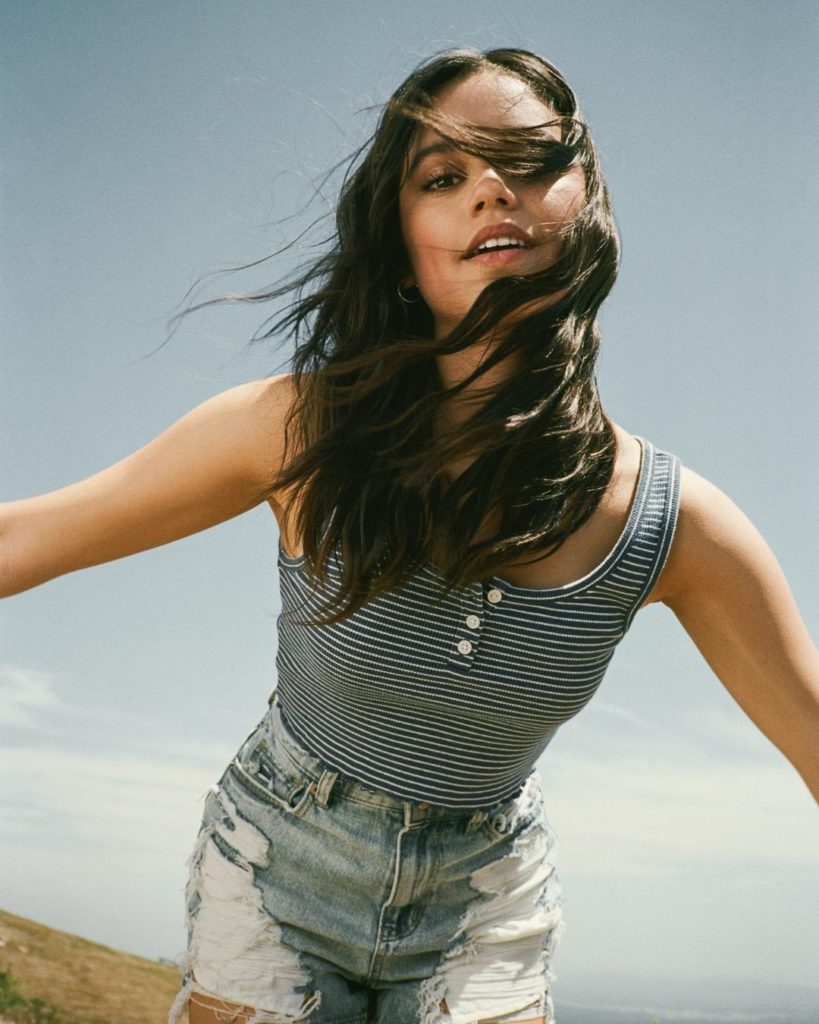 Jenna leaning forward, wind blowing through her hair, wearing a blue-white striped top and worn jeans.