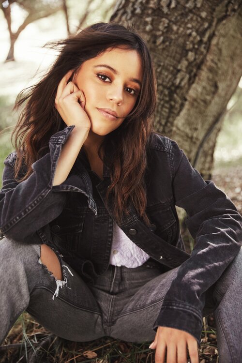 Jenna sitting in front of a tree, wearing a black jeans jacket over a  white shirt and matching dark jeans